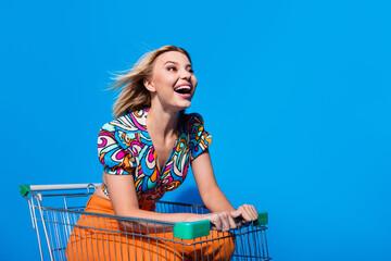 Wall Mural - Full length photo of overjoyed funky girl ride fast on shopping cart staring at empty space promo isolated on blue color background