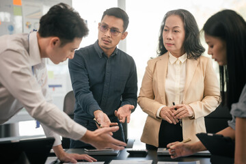 Wall Mural - diverse group of Asian professionals, including middle-aged and mature individuals, gathered around a table in a business setting, discussing documents with focused attention.