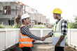 © R Photography - two construction workers, one Asian, shaking hands on a building site, possibly indicating agreement or completion of a task, with a modern building in the background.