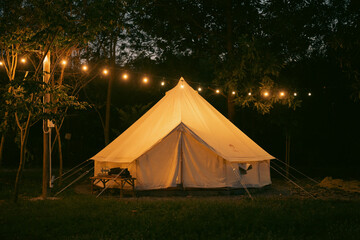 night and warm light exposure of an illuminated bell tent surrounded by trees and a small camp fire 