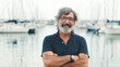 Happy male seigneur in glasses crossing his arms while standing in the port and looking at the camera on yachts background, camera is moving closer