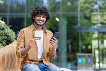 Portrait of a smiling young Indian man sitting outside on a bench, holding a credit card and a phone, looking at the camera