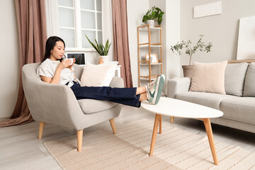 Sticker - Young Asian woman drinking coffee in armchair at home