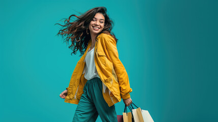 Stylish woman in a yellow coat walking and carrying shopping bags, against a solid teal background.