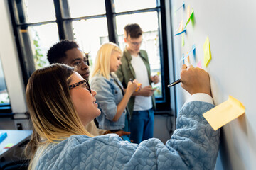 Diverse group of young business people working in office using colorful sticky notes on wall to organize their ideas.