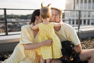 Wall Mural - Happy parents wearing yellow outfits sitting next to each other while little daughter hugging and covering faces. Caucasian spouses showing love in family while spending time with kid and dog outside.