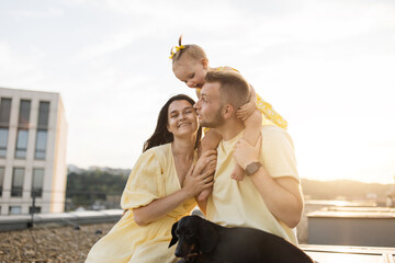 Wall Mural - Happy family of three with dog enjoying beautiful sunset on rooftop. Handsome father holding little daughter on shoulders and they both smiling to each other while mother sitting near with pet.