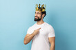 Portrait of proud man with beard wearing white T-shirt and golden crown and pointing himself, looking at camera with smile, superior privileged status. Indoor studio shot isolated on blue background.