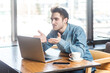 © khosrork - Portrait of positive romantic young man freelancer in blue jeans shirt working on laptop, having video call, sanding air kisses over palms. Indoor shot near big window, cafe background.