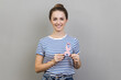 Portrait of woman smiling to camera and holding pink ribbon, symbol of breast cancer awareness, diagnostics and prevention of oncological disease. Indoor studio shot isolated on gray background.