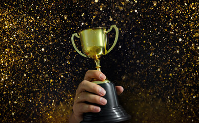 Wall Mural - man's hand holding a golden trophy with golden glitter sparkling around it, competition concept for 