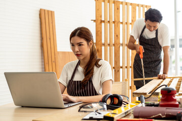 Two woodworker; one focused on crafting with wood while the other uses a laptop for design insights.