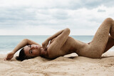Gorgeous young woman lying in the sand, her body artfully covered with grains of sand, on a beach