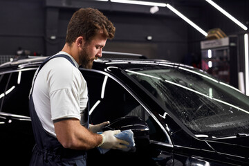 Canvas Print - Young auto mechanic polishing or cleaning black automobile with rag. Manual car washing. In Auto service, man in uniform and gloves cleaning vehicle