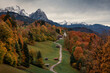 Bavarian Alps with church of Wamberg in Garmisch-Partenkirchen during autumn, snow-covered mountains in the background, dramatic cloudy sky, colored leaves and trees and huts and road in foreground