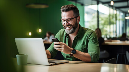 Wall Mural - Cheerful man with a beard and glasses working on a laptop at a wooden desk