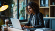 © Studio Nova - Young woman seated and smiling at the camera, with a laptop in front of her and a blurred office environment in the background