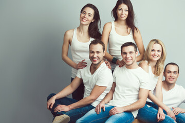 Happy together concept. Group portrait of healthy boys & girls in white t-shirts, sleeveless shirts and blue jeans standing, sitting, posing over gray background. Urban street style. Copy-space