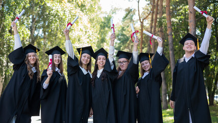 Wall Mural - Row of young students in graduation gowns outdoors showing off their diplomas. 