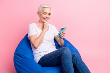 Poster - Full body photo of minded person dressed stylish t-shirt denim pants sit on pouf look at smartphone isolated on pink color background