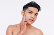 © Mdv Edwards - A young asian man applies exfoliating facial scrub cream to his face using his fingers. Skin care and hygiene concepts. Isolated on a white backdrop.