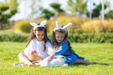 Wall Mural - Two little girls with a bunny rabbit have an Easter at green grass background. Two children wearing Bunny ears find and pick up multicolored egg on Easter egg hunt in garden.