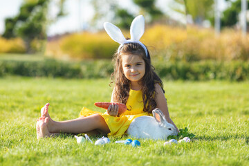 Wall Mural - Happy little girl with a bunny rabbit have an Easter at green grass background. Child wearing Bunny ears find and pick up multicolored egg on Easter egg hunt in garden.