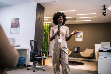 Young business woman lecturing her multiethnic colleagues. Having a speech while holding a microphone.