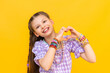 © Юлия Дьякова - The little girl shows her heart with her fingers and smiles happily. The child put on a lot of beaded bracelets. Beading for children. Yellow isolated background.