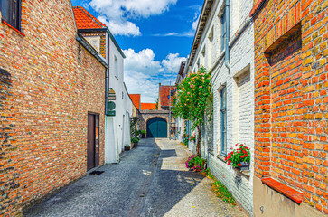 Empty narrow cobblestone dead end quiet street, buildings with flowers on brick walls in summer sunny day, Brugge city historic centre, Bruges old town quarter district, Flemish Region, Belgium