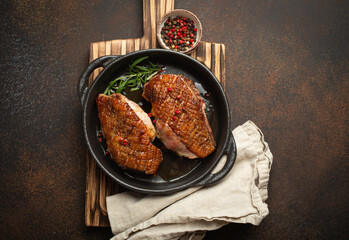 Wall Mural - Two roasted duck breast fillets with crispy skin, with pepper and rosemary, top view in black cast iron pan with knife, dark brown concrete rustic background.