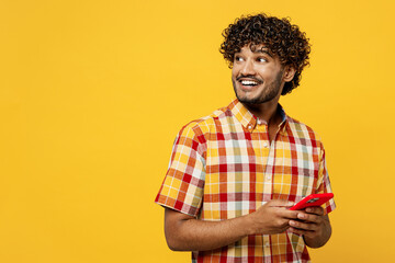 Wall Mural - Side view young smiling happy Indian man wears shirt casual clothes hold in hand use mobile cell phone look aside on area isolated on plain yellow color background studio portrait. Lifestyle concept.