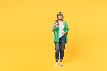 Wall Mural - Elderly blonde smiling happy fun woman 50s years old wears green shirt glasses casual clothes hold in hand use mobile cell phone typing message isolated on plain yellow background. Lifestyle concept.