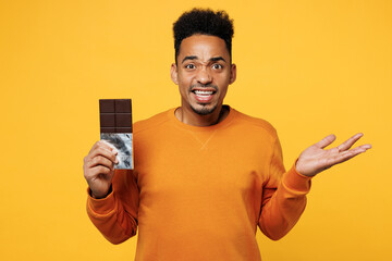 Wall Mural - Young confused sad man he wear orange sweatshirt casual clothes hold eat bar of chocolate spread hand isolated on plain yellow background. Proper nutrition healthy fast food unhealthy choice concept.