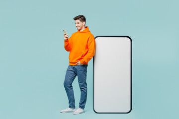 Canvas Print - Full body young man he wearing orange hoody casual clothes stand near big huge blank screen mobile cell phone with area use smartphone isolated on plain blue cyan color background. Lifestyle concept.