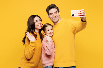 Wall Mural - Young parents mom dad with child kid girl 7-8 years old wear pink knitted sweater casual clothes do selfie shot on mobile cell phone waving hand isolated on plain yellow background Family day concept