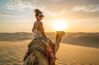 © Jose Luis Stephens - A young woman enjoys a camel ride through sweeping desert dunes as the sun sets, casting warm, golden hues across the sandy scenery