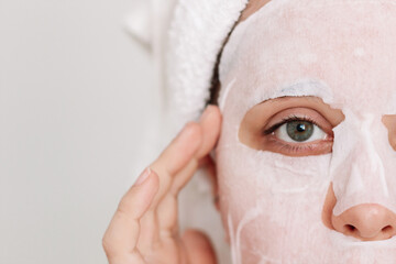 Wall Mural - Cropped shot of a young caucasian woman with a white towel on her head after a shower putting a fabric cosmetic moisturizing mask on her face on a light background. Skin care, cosmetology