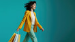 Stylish woman in a yellow coat walking and carrying shopping bags, against a solid teal background.