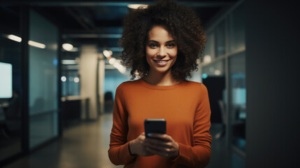 Wall Mural - Smiling African American woman wearing a red sweater, enjoying a moment while using her smartphone in a modern office environment.