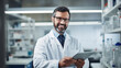 © MP Studio - Smiling male scientist with curly hair and glasses is holding a tablet and standing in a modern laboratory