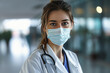 mujer doctora portando mascarilla quirúrgica, bata blanca y estetoscopio sobre fondo desenfocado de pasillo de hospital