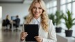 © Tom - A bright and modern office setting featuring a cheerful blonde businesswoman holding a black tablet, with colleagues and indoor plants in the background.