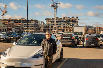 green economy: a senior citizen ( man with hat and beard) stands beside a new white electric vehicle in mall parking lot with high rise construction  in background.