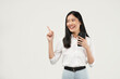 © Illhamt - Photo of young asian girl shopping on mobile app, holding phone and pointing finger left at empty copy space, showing promo text, standing with smartphone against white background