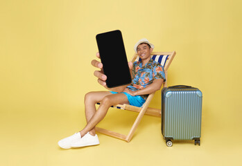 Wall Mural - Young asian man wear summer clothes sit in deckchair showing mockup mobile cell phone app booking hotel isolated on yellow background.