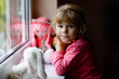 Cute toddler girl sitting by window and looking out on rainy day. Dreaming child with doll and soft toy feeling happy. Self isolation concept during corona virus pandemic time. Lonely kid.