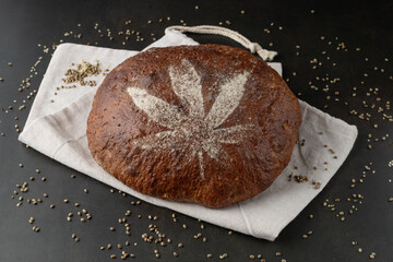 Wall Mural - Fresh baked loaf of spelt bread with crushed hemp seeds. Bread decorated with cannabis leaf made of flour on hemp cloth bag on brown grunge background.