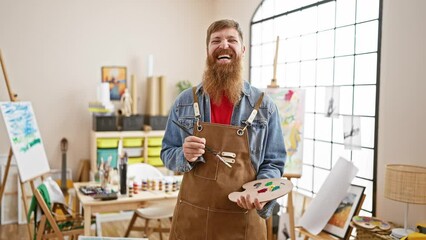 Sticker - Confident young redhead man artist paintbrush in hand, smiling joyfully at an indoor art studio