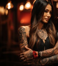 A woman with tattoos in various styles poses with her arms crossed in an elegant black top and red bracelet, in front of a dimly lit bar.